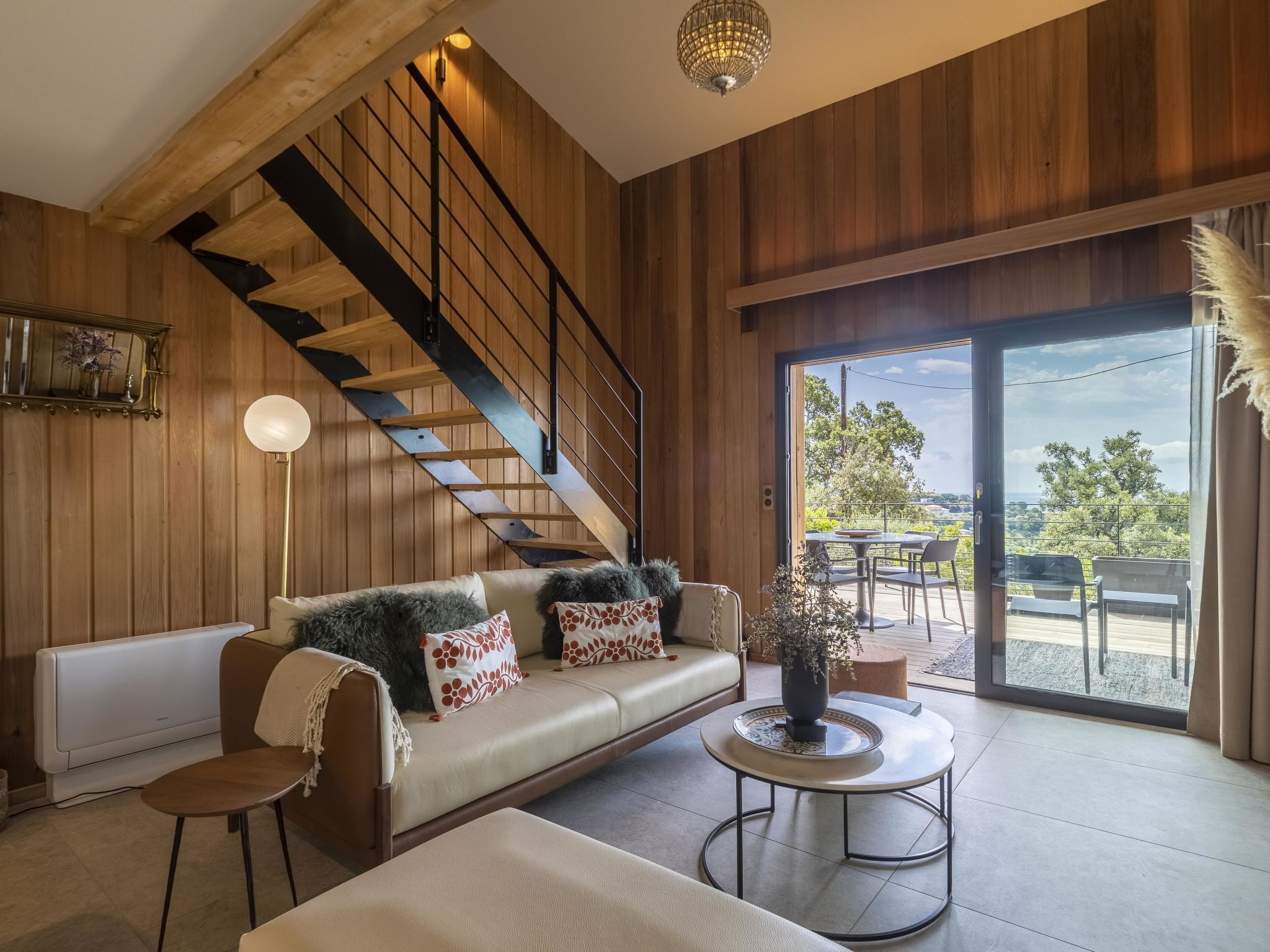 Uma sala de estar moderna com escada de madeira, um sofá com almofadas estampadas, uma mesa de centro redonda e vista para o exterior através de uma porta de vidro.