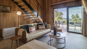 Un salon moderne avec un escalier en bois, un canapé agrémenté de coussins à motifs, une table basse ronde et une vue sur l’extérieur grâce à une porte vitrée.