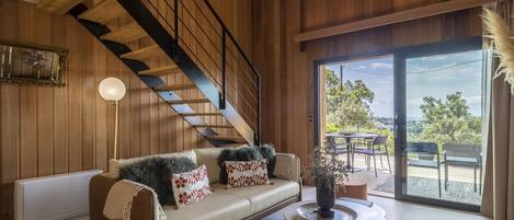 Una sala de estar moderna con una escalera de madera, un sofá con cojines estampados, una mesa redonda de centro y vistas al exterior a través de un ventanal.