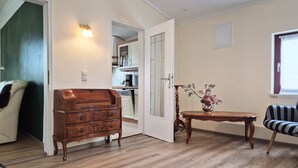 Une pièce avec une commode en bois, une petite table sur laquelle est posé un vase, une chaise à rayures et, au fond, un coin cuisine.
