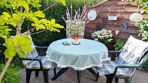 Une table ronde recouverte d’une nappe au crochet, deux chaises avec des coussins à carreaux et un pot de fleurs orné de fleurs roses.