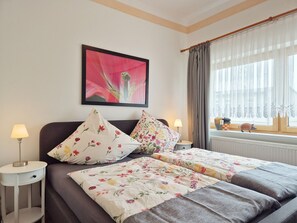 Une chambre d’hôtel avec un lit, deux tables de chevet, une lampe, une fenêtre avec des rideaux, un objet décoratif sur le rebord de la fenêtre et un tableau encadré au mur.