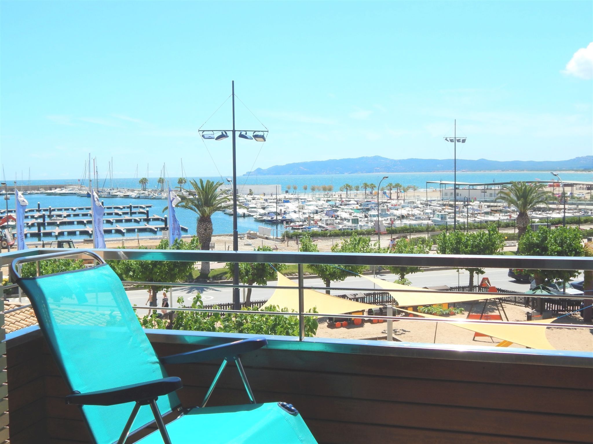 A balcony with a turquoise chair overlooking a marina with boats and a beach in the background.