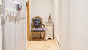 Un couloir étroit doté d’un parquet en bois disposé en point de Hongrie, d’un fauteuil bleu recouvert de tissu, d’une petite commode blanche et d’une porte avec un interrupteur.