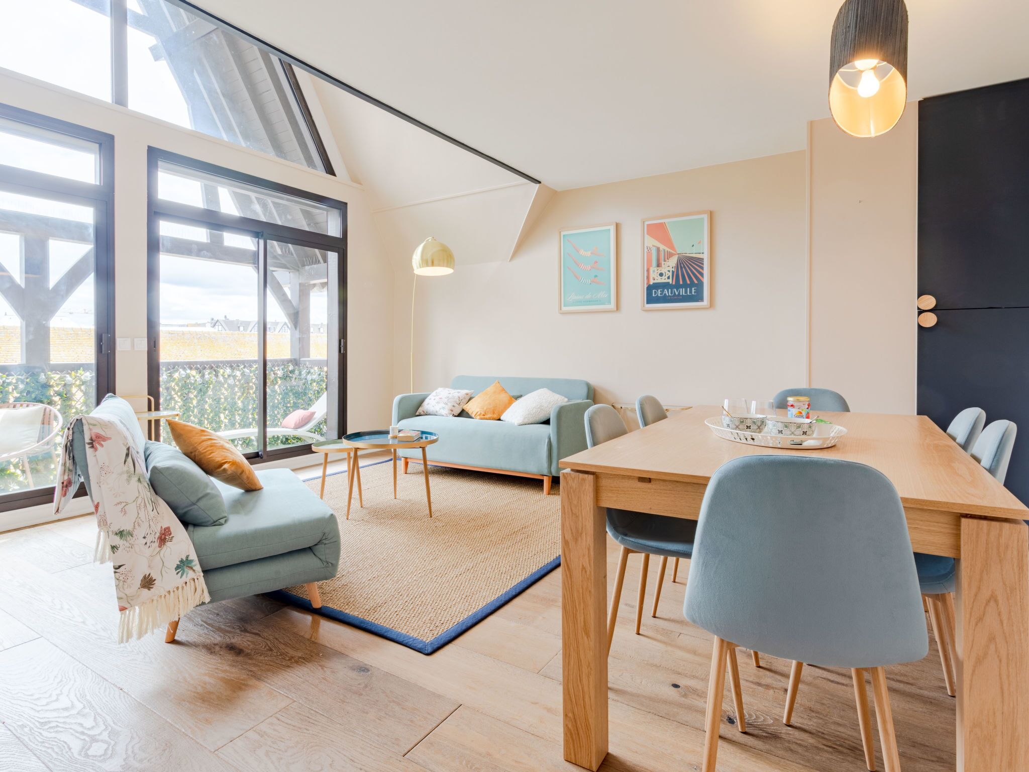 Een moderne woonkamer met een bank, salontafel en een eettafel. Twee ingelijste foto’s hangen aan de muur en een groot raam zorgt voor natuurlijk licht.