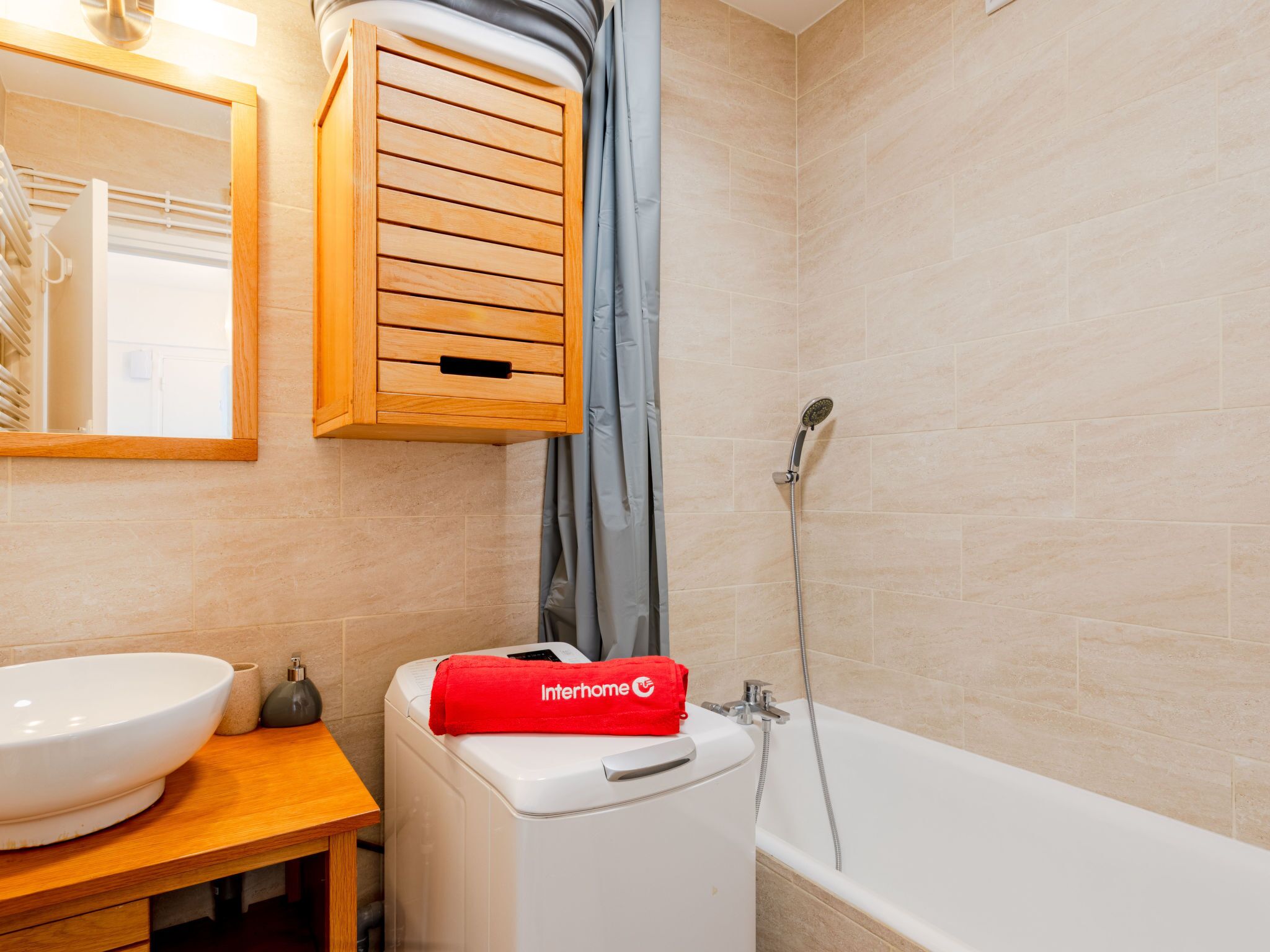 A bathroom with a white sink, a wooden cabinet, a shower with a handheld showerhead, and a red Interhome towel on a washing machine.