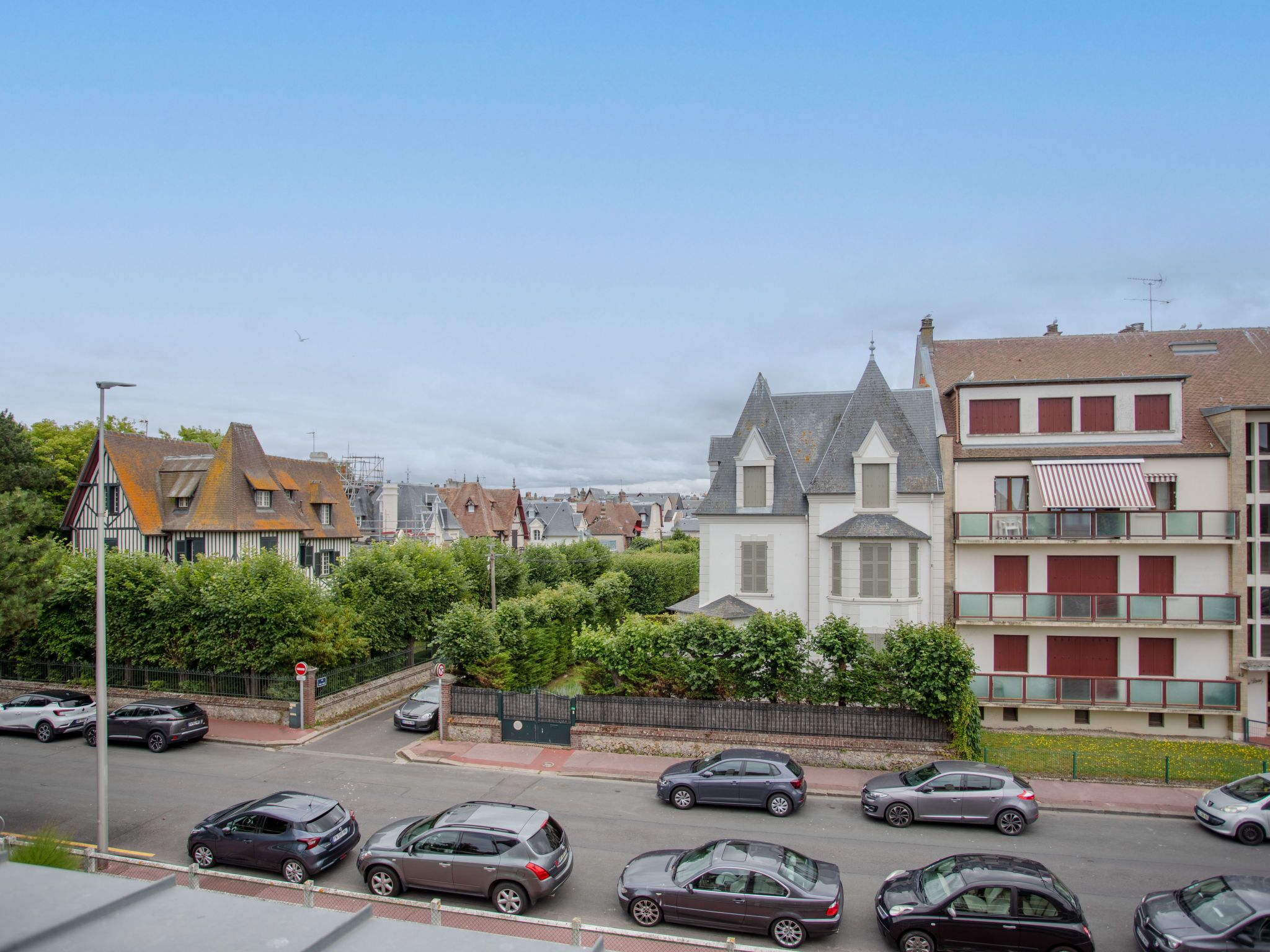 A street view with parked cars, a mix of residential buildings, and a prominent house with a gabled roof.
