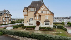 Une grande maison de style Tudor à deux étages, dotée d’une tourelle imposante et de larges baies vitrées, située dans une zone côtière, avec une marina visible au loin.