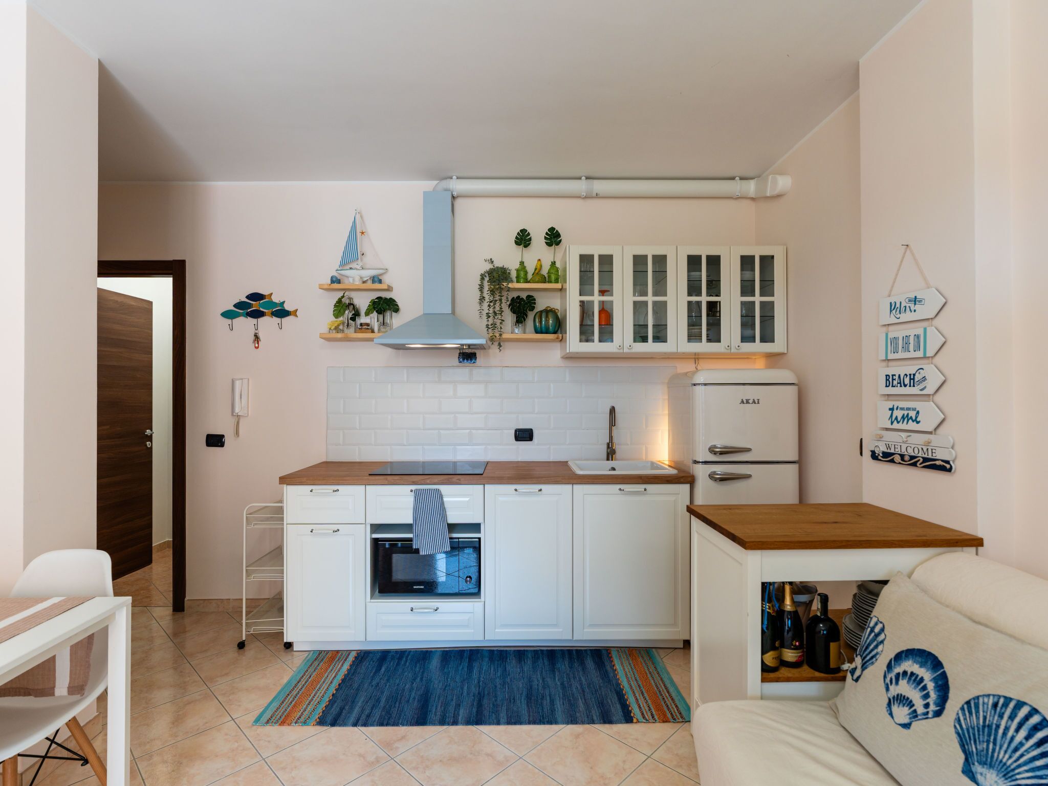 A compact kitchen with white cabinets, a built-in oven, and a compact refrigerator. There is a wooden countertop, a blue rug, and a small seating area with a sofa and a coffee table.
