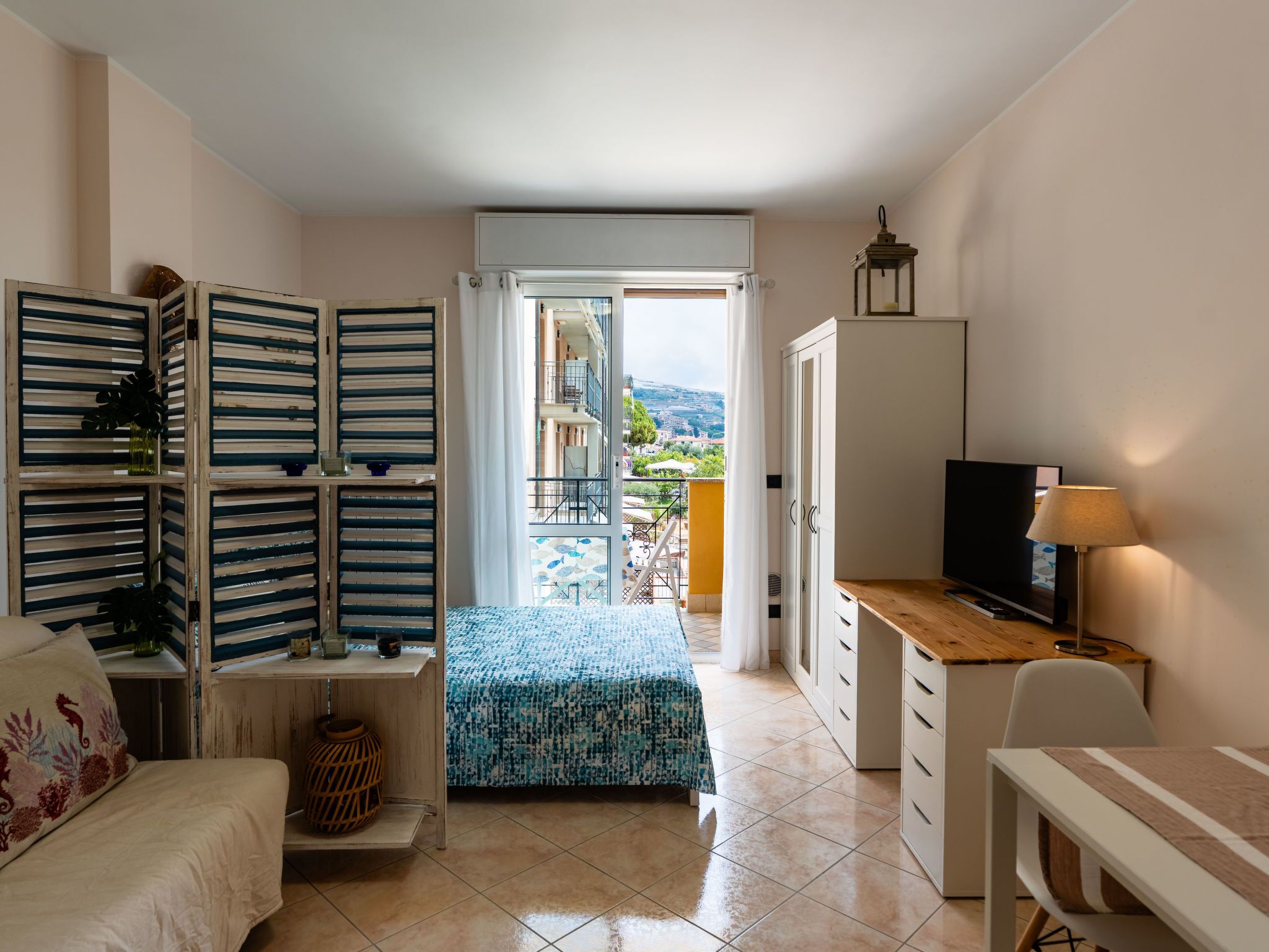 A compact room with a bed, a desk, a TV, and a balcony with a view of the city.