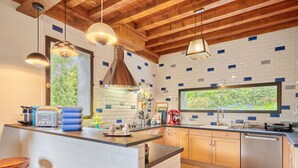 A modern kitchen with a central island, wooden ceiling beams, and a large window overlooking greenery.