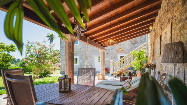 Un patio al aire libre con mesa y sillas de madera, paredes de piedra y vista a un edificio al fondo.