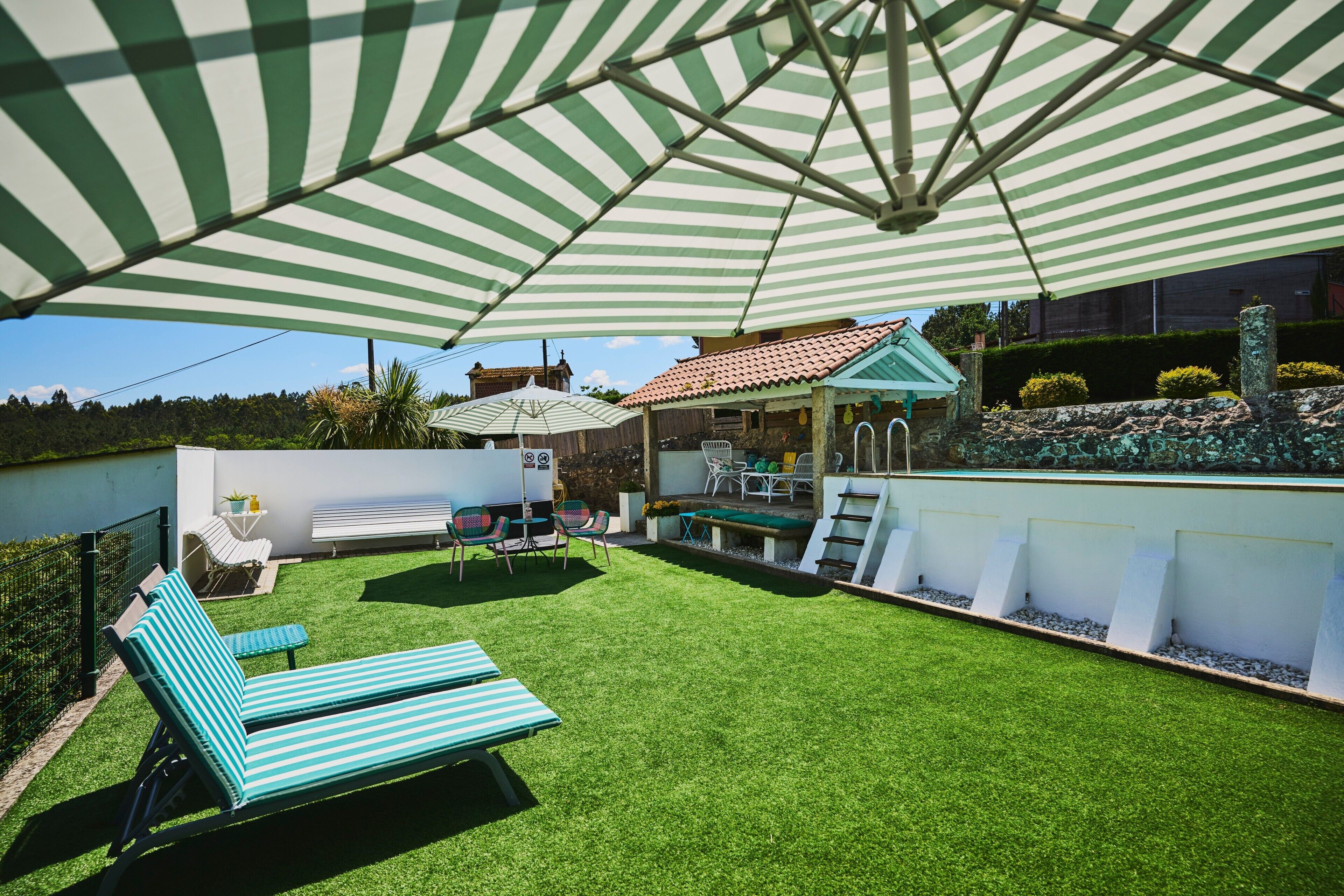 Ein Garten mit Kunstgrasanlage, einem gestreiften Sonnenschirm, Liegestühlen und einer kleinen Terrasse.