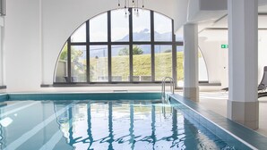 An indoor swimming pool with clear blue water, large windows offering a view of greenery and mountains, and a metal ladder for entering the pool. - La Siala Apt.66 by Interhome (Falera)