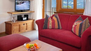 A living room with a red sofa, a wooden coffee table with a fruit bowl, a TV on a wooden shelf, and a window with a mountain view. - La Siala Apt.66 by Interhome (Falera)