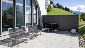 A modern outdoor seating area with metal chairs and a table, large windows, and a view of a hillside with a house. - La Siala Apt.35 by Interhome (Falera)