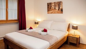 A wooden bed with white bedding, two bedside tables with lamps, a window with sheer curtains, and a painting of a forest on the wall. - La Siala Apt.35 by Interhome (Falera)