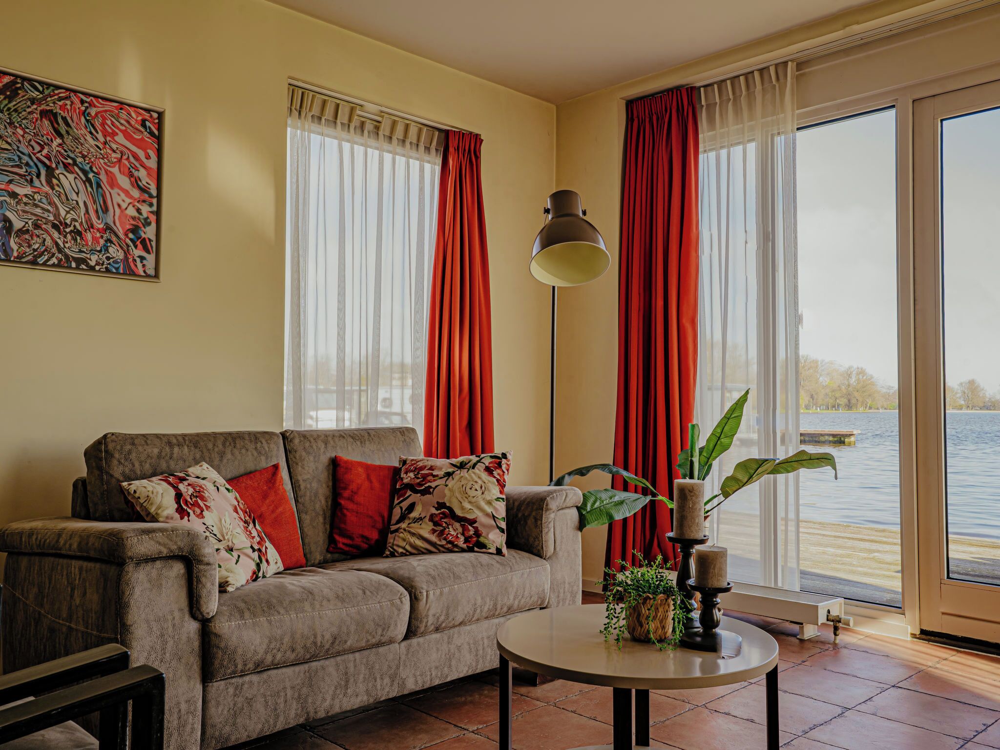 A living room with a sofa, a table, and a view of a body of water through large windows with red curtains.