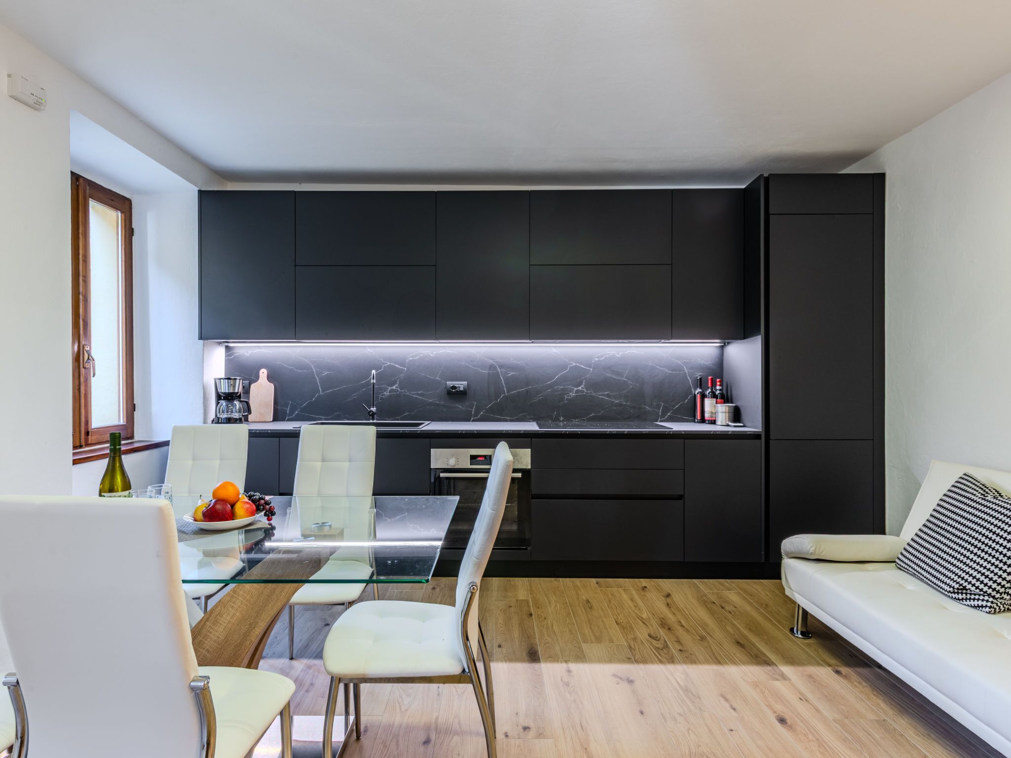 A modern kitchen with dark cabinets, a marble backsplash, and a glass dining table with chairs.