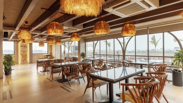 A spacious indoor dining area with wooden tables and chairs, large windows offering views of palm trees and buildings, and hanging thatched lamps. - Hotel Royal Bagh - Opposite City Park (Jaipur)