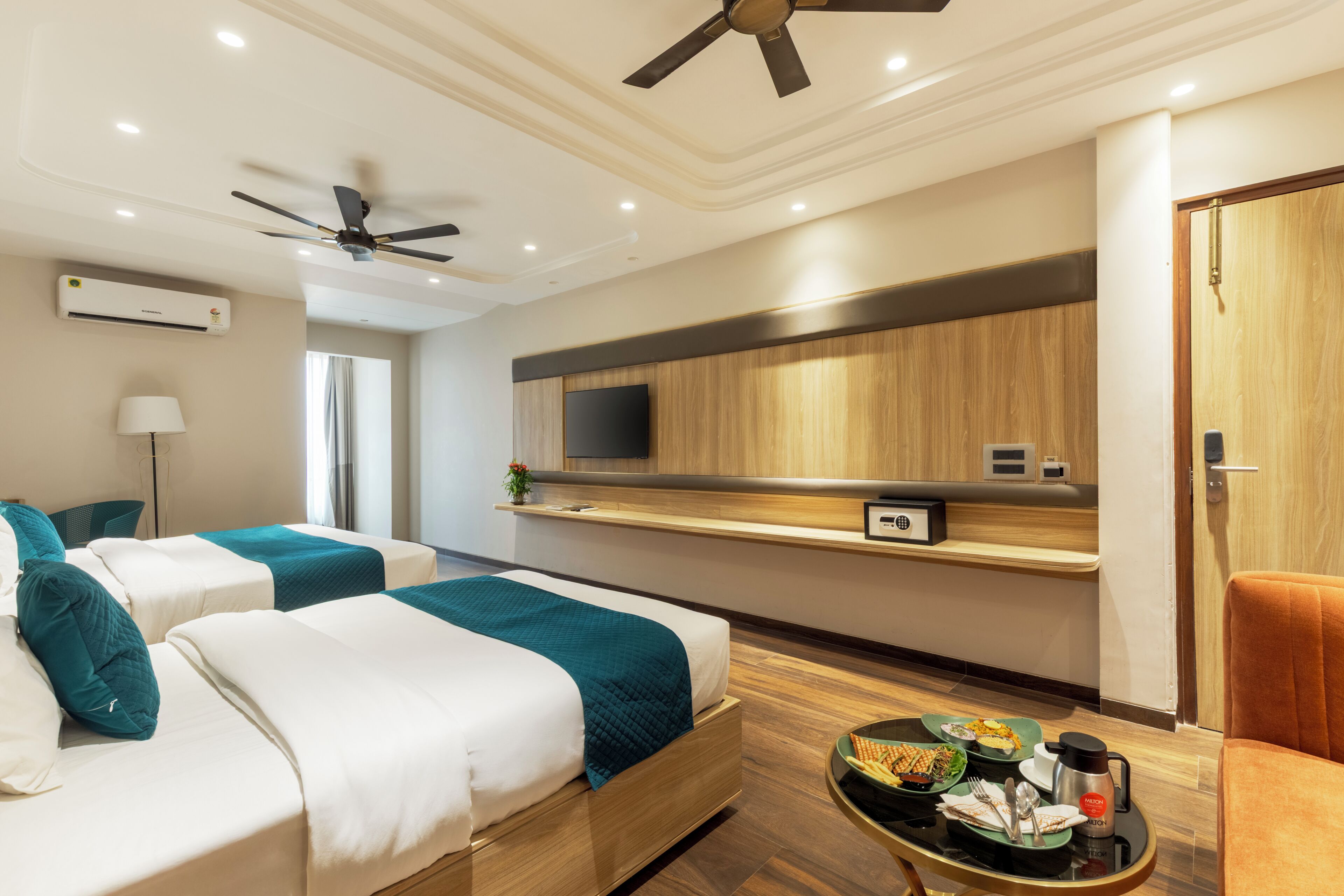 A hotel room with two beds, a TV mounted on the wall, a ceiling fan, and a small table with breakfast items.