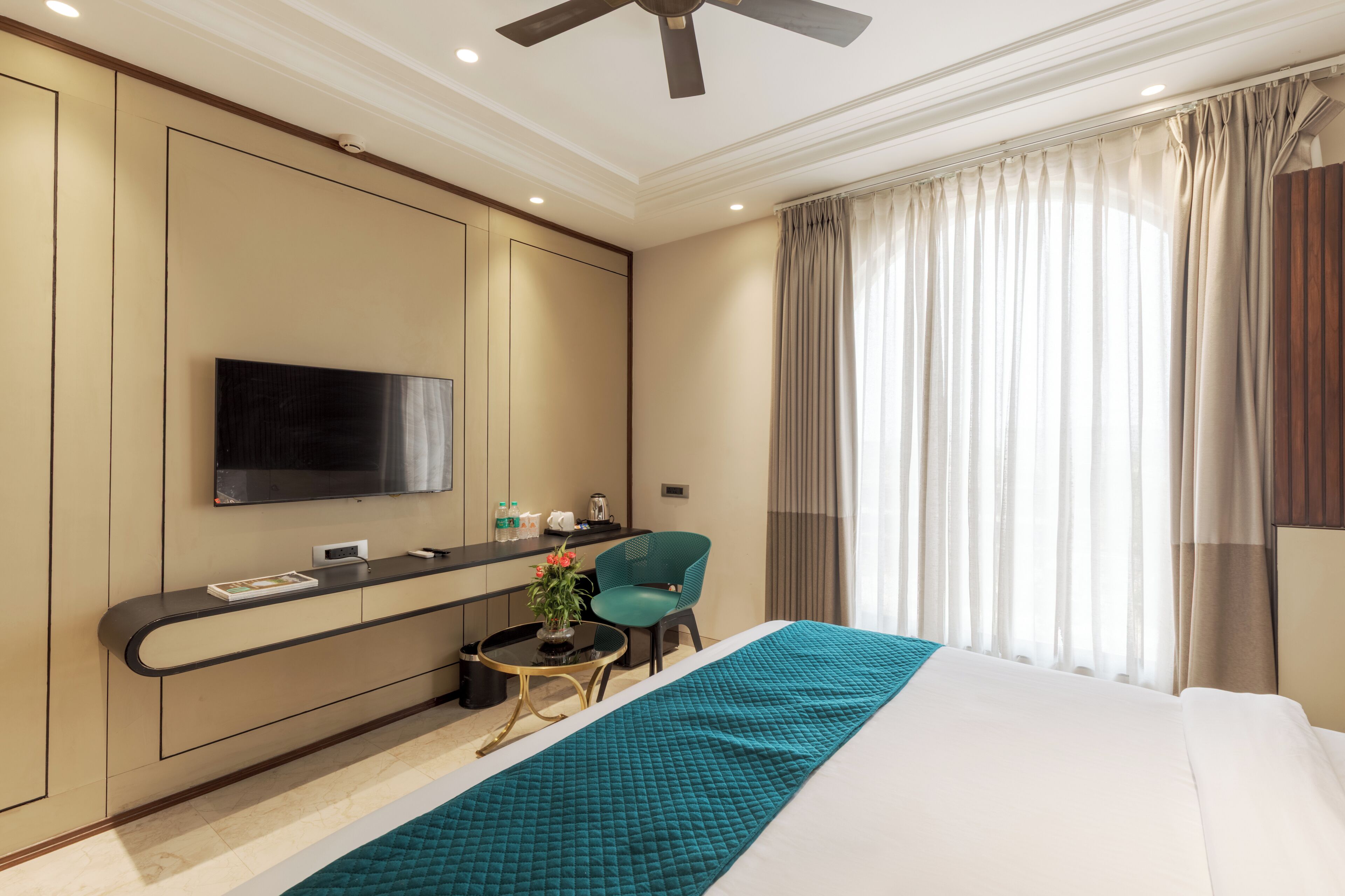 A hotel room with a bed, a desk with a flat-screen TV, a chair, a small table with a plant, and a window with curtains.