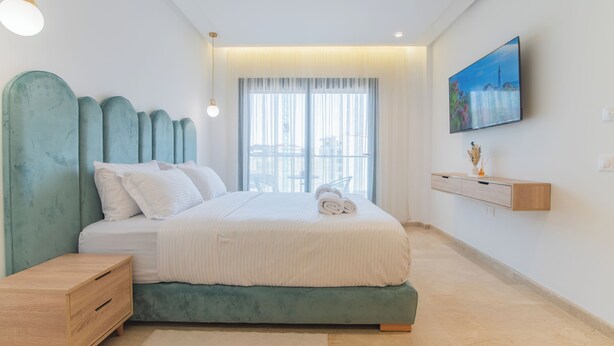 Une chambre à coucher moderne avec un grand lit, une table de chevet, un téléviseur fixé au mur et une fenêtre ornée de rideaux légers.