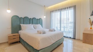 Une chambre moderne avec une tête de lit turquoise, un lit soigneusement fait, une table de chevet en bois et une grande fenêtre ornée de rideaux légers.