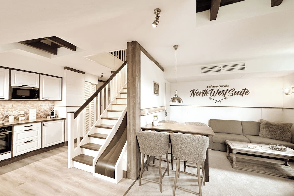 A modern kitchen with a microwave, stove, and cabinets. There is a staircase leading to a loft area. A dining table with chairs is in the center. A sofa and a coffee table are in the living area. A sign on the wall reads "welcome to the NorthWest Suite".
