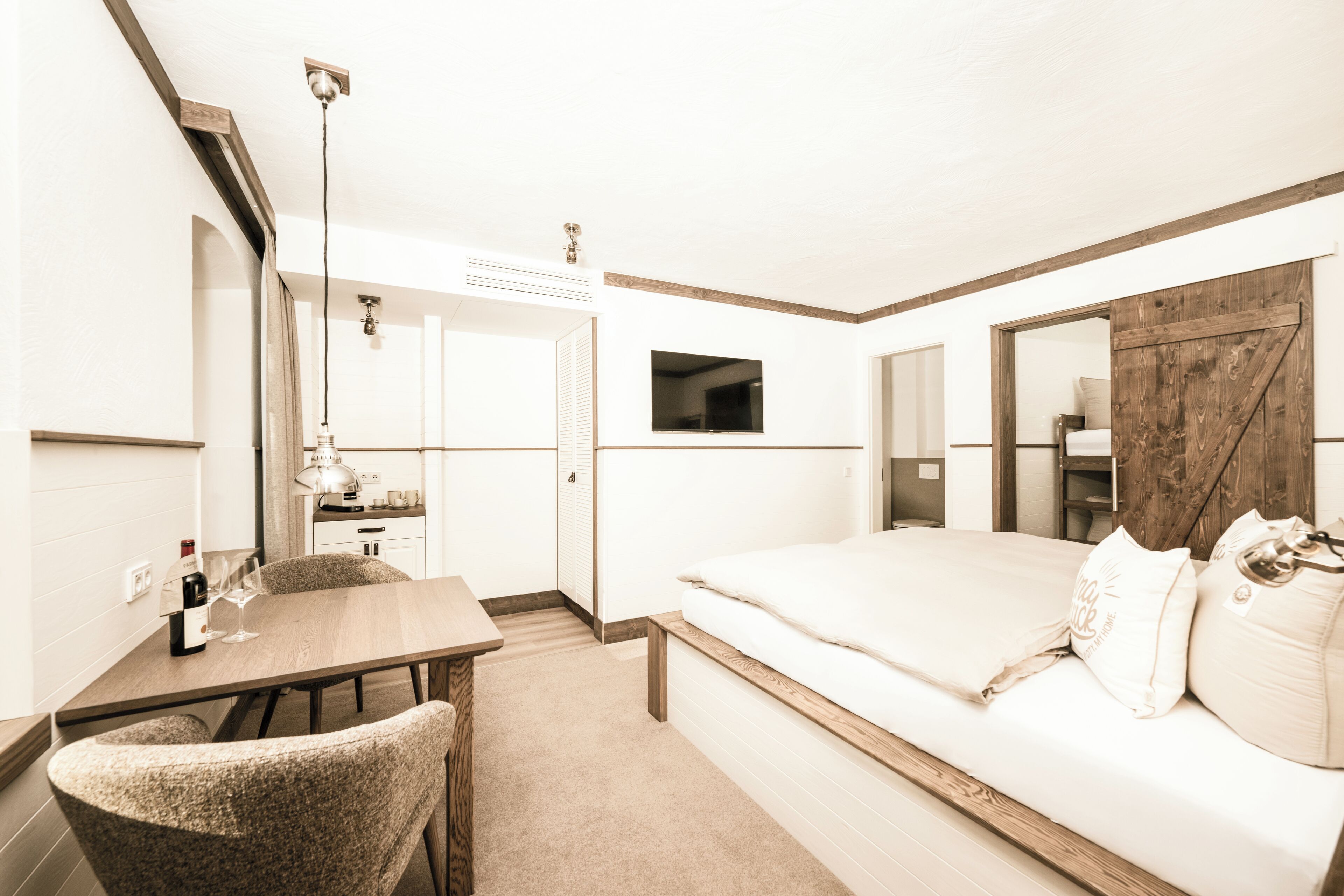 Une chambre d’hôtel avec un lit, un bureau et une chaise, une petite table avec une bouteille et des verres, et un téléviseur fixé au mur.