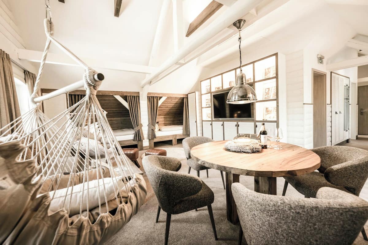 A modern room with a round wooden table, six grey upholstered chairs, a macramé hanging chair, a built-in TV shelf, and a wooden ceiling.