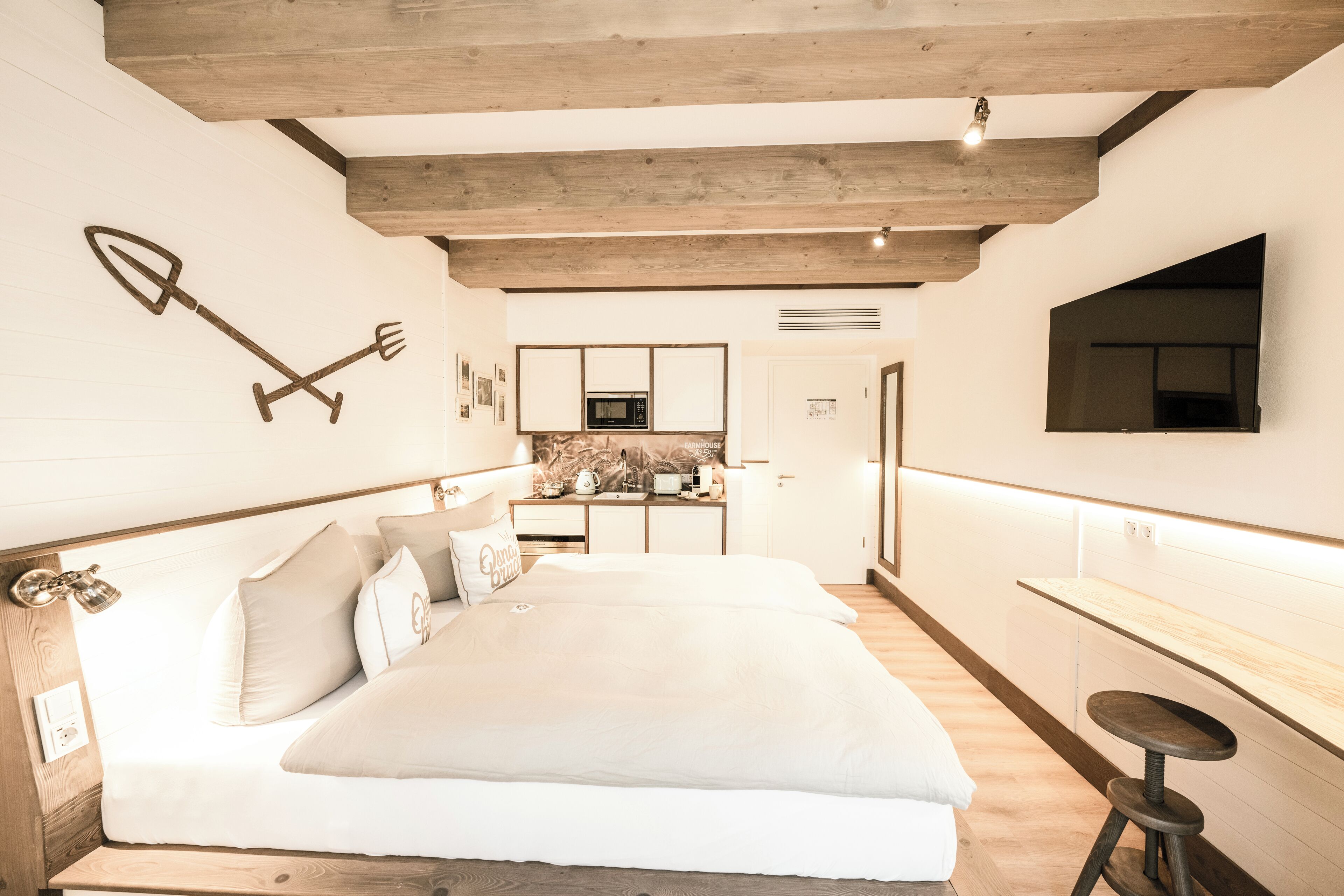 A bedroom with a large bed, wooden beams, a kitchen area, and a TV mounted on the wall.
