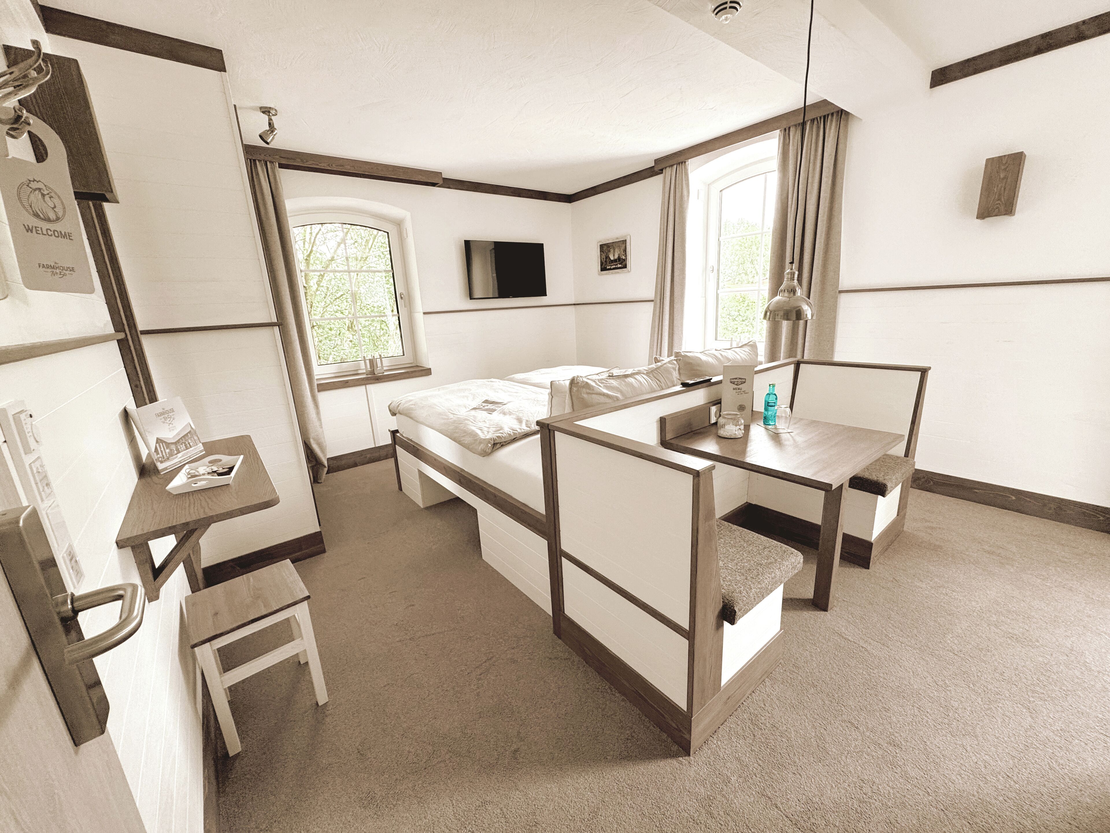 A compact hotel room with a bed, a desk, a chair, a small table, a TV, and a window.