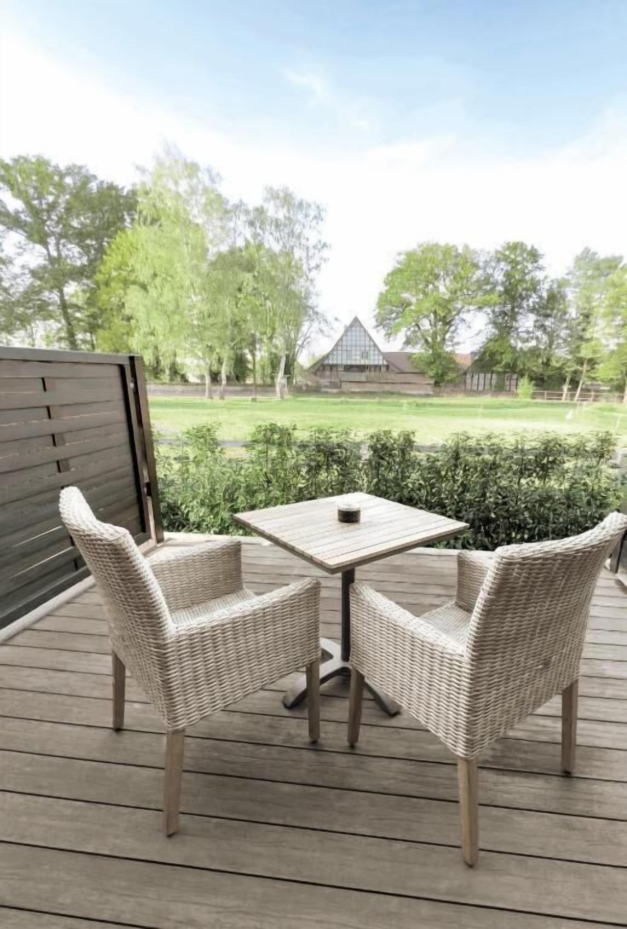 A deck with wicker chairs and a table, overlooking a grassy area with trees and a building in the background.