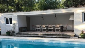 Een overdekte eethoek buiten, met een zwembad, stoelen en hangende lichten.