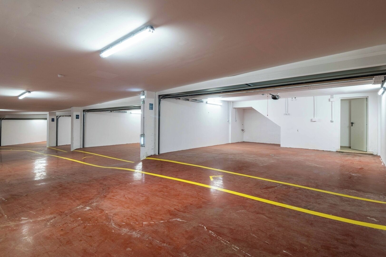 A spacious indoor parking garage with multiple parking bays, marked by yellow lines, and white walls.