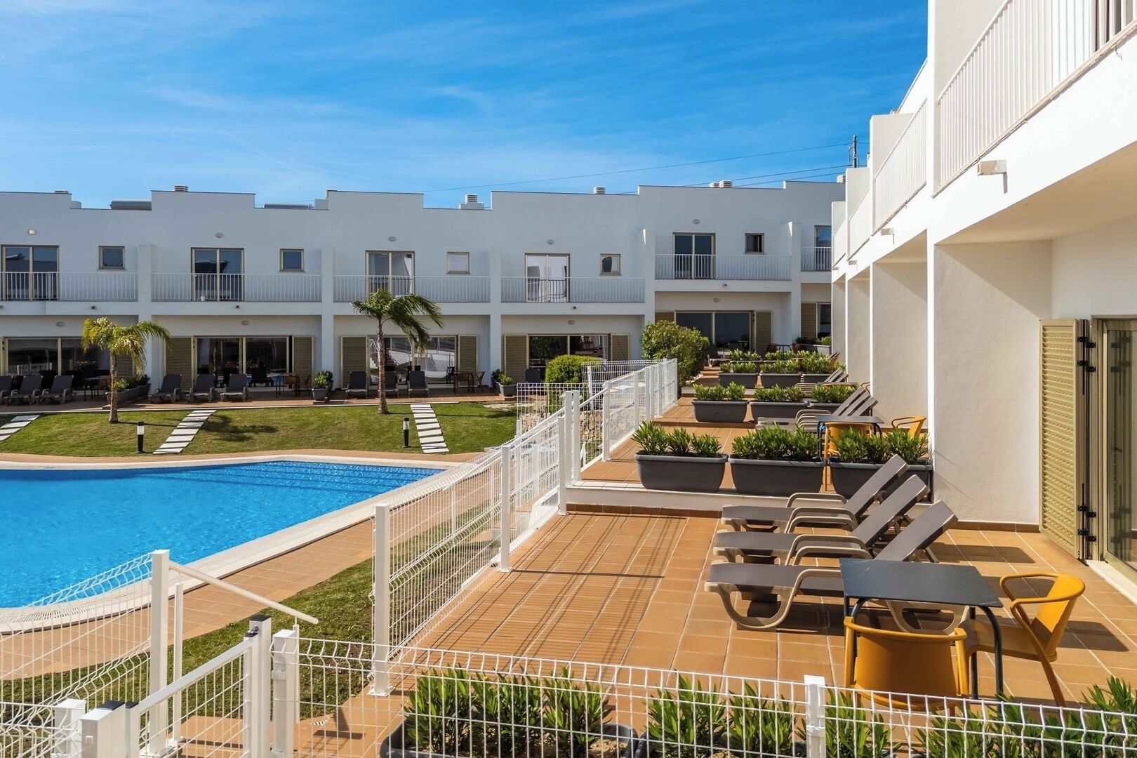 A rooftop pool area with lounge chairs and potted plants, surrounded by a white building with balconies.