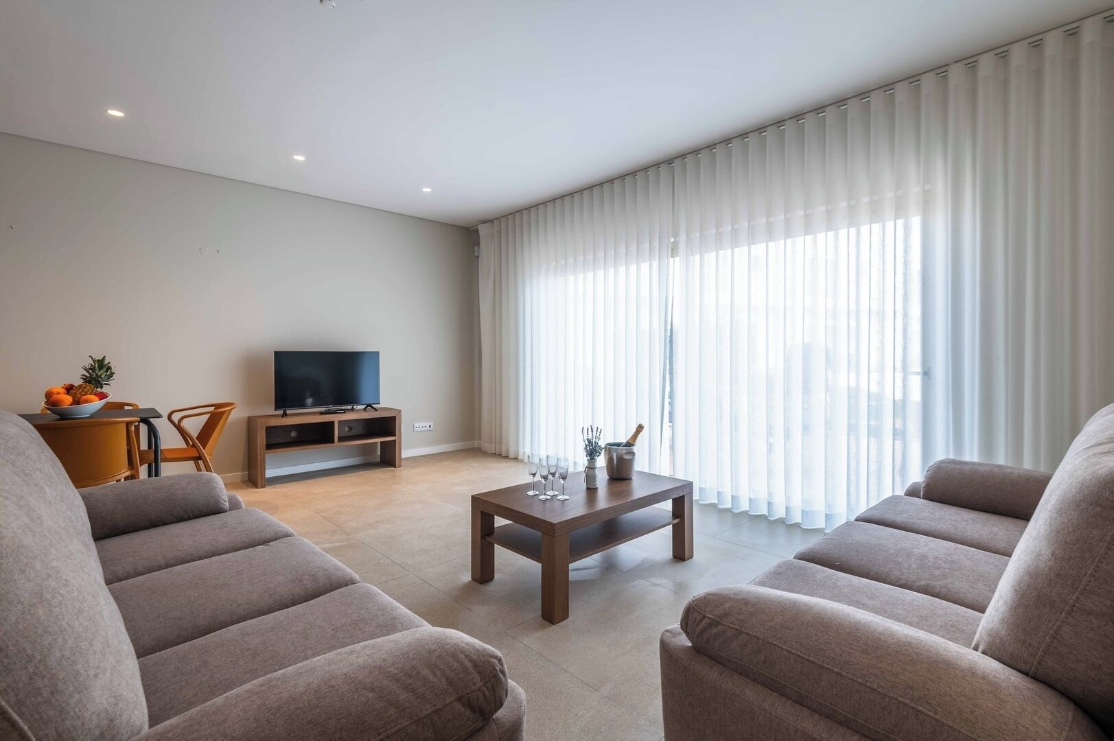 A modern living room with two sofas, a coffee table, a TV, and a dining area with a fruit bowl.