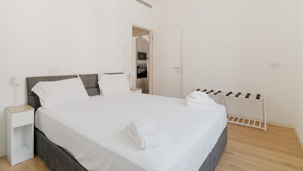 A neatly made bed with white linens, a bedside table with a lamp, and a white drying rack. - GuestHost - Cozy Apartment X4 in Navigli District (Milan)