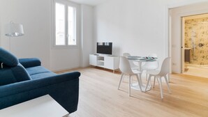 A modern living room with a blue sofa, a white dining table with chairs, a TV on a white cabinet, and a large window. - GuestHost - Cozy Apartment X4 in Navigli District (Milan)