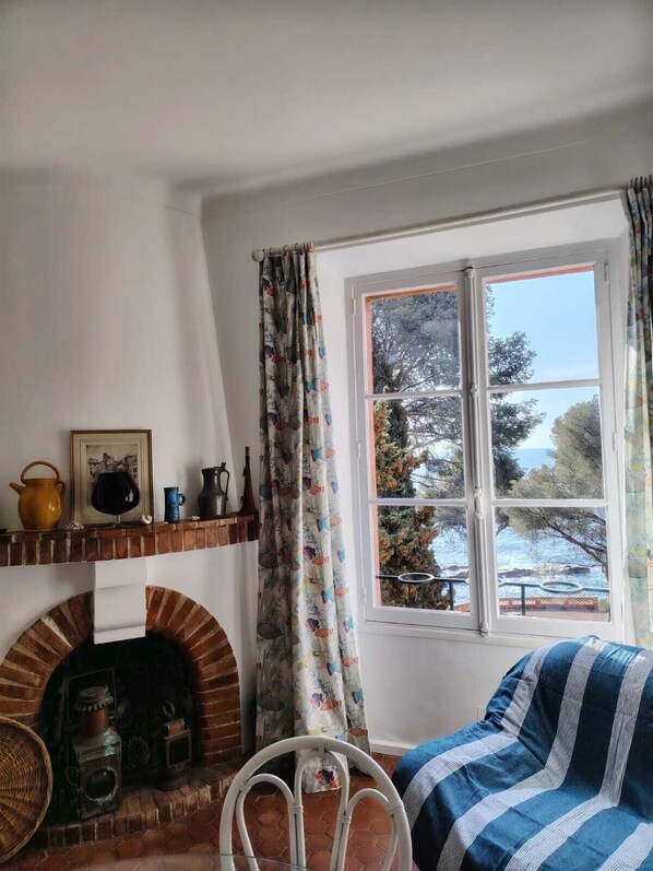 A cozy room with a brick fireplace, a window with a view of trees and the sea, and a blue-striped bed. - Waterfront 3-room apartment (Saint-Raphaël)