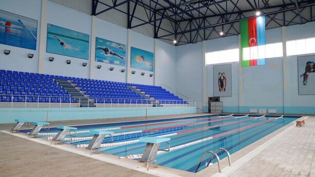 En swimmingpool med flere baner, blå siddepladser på tilskuerpladserne, og et flag på væggen.
