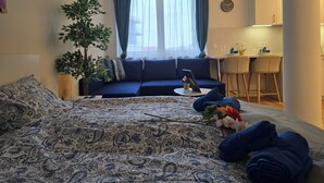 A cozy room with a bed, a blue sofa, a small table with a laptop, a potted plant, and a view of a kitchen area.