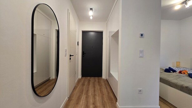 A hallway with a wooden floor, a black door at the end, and a bed with a basket and flowers on the right.