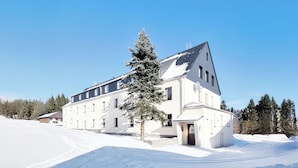 A large, white building with a sloped roof, surrounded by snow and trees. - Hotel Erlebnis Wald (Johanngeorgenstadt)