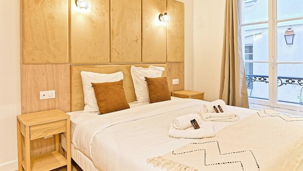 A hotel room with a large bed, wooden headboard, bedside lamps, a window with a view, and a neatly made bed with towels and toiletries. - Magnificent Air-conditioned Flat Galeries Lafayette (Paris)