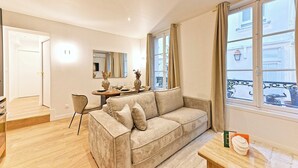 A living room with a beige sofa, a dining table, and a window with curtains. - Magnificent Air-conditioned Flat Galeries Lafayette (Paris)