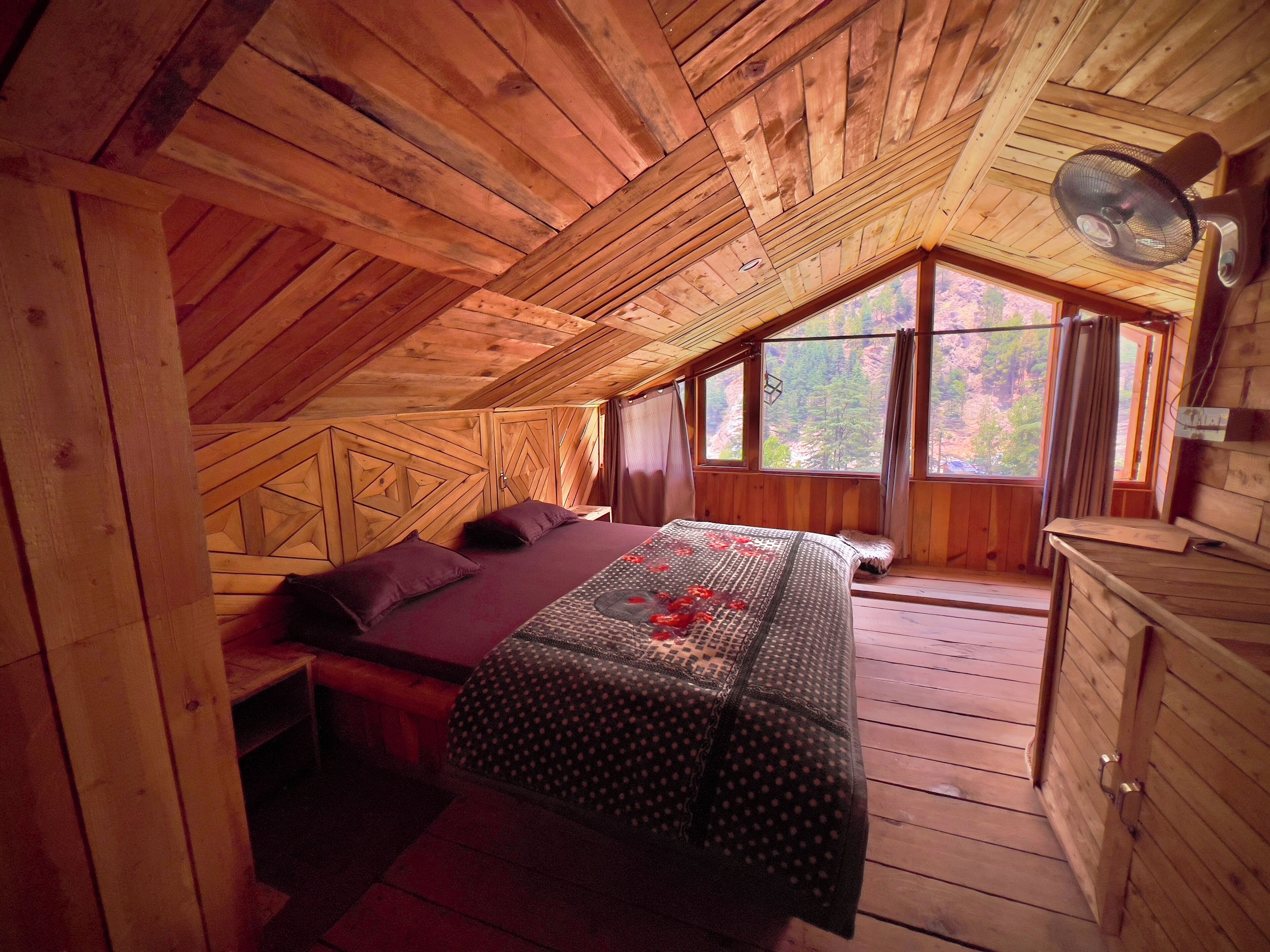 A cozy attic bedroom with a wooden ceiling, a bed with a patterned bedspread, a dresser, and a window with a view of trees.