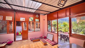 A room with a wooden floor, a sofa, a table, and large windows offering a view of greenery outside. - Royal Star Kasol (Kasol)