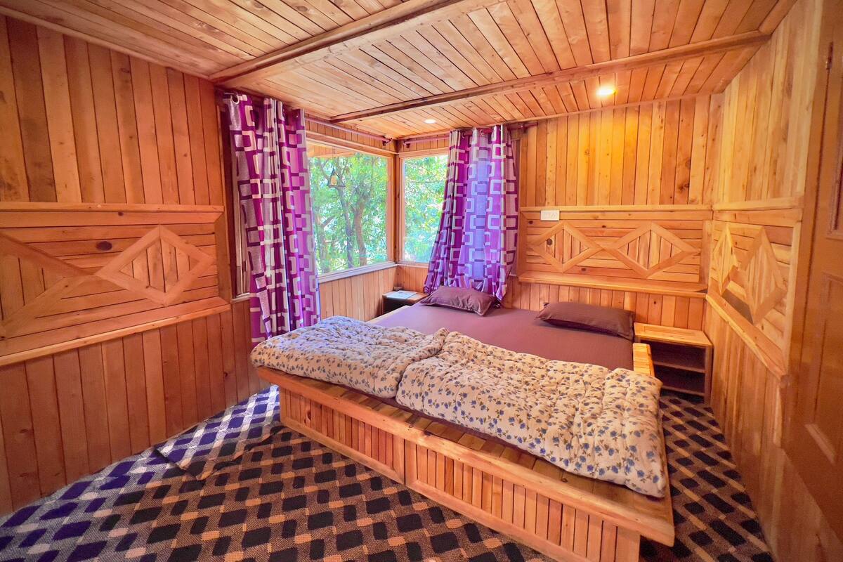 A wooden cabin room with a bed, curtains, and a window with a view of trees.